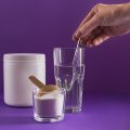 Glass with collagen dissolved in water and collagen protein powder on purple background. Woman's hand holds a spoon. Healthy lifestyle concept.