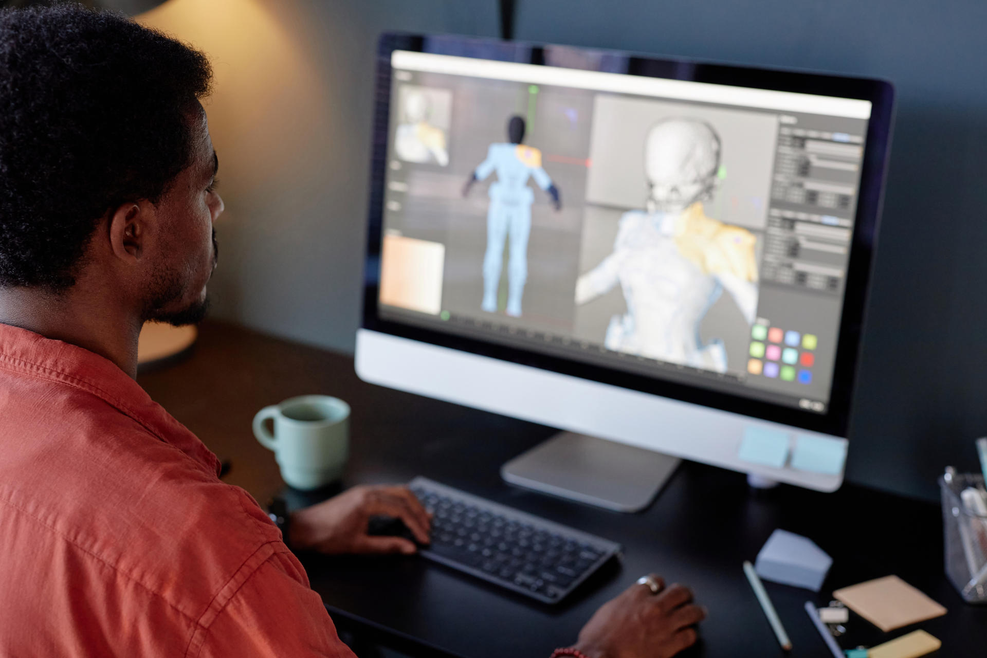 Individual sitting at desk while handling a 3D modeling project on computer monitor. Background features various design tools on the screen and desk accessories