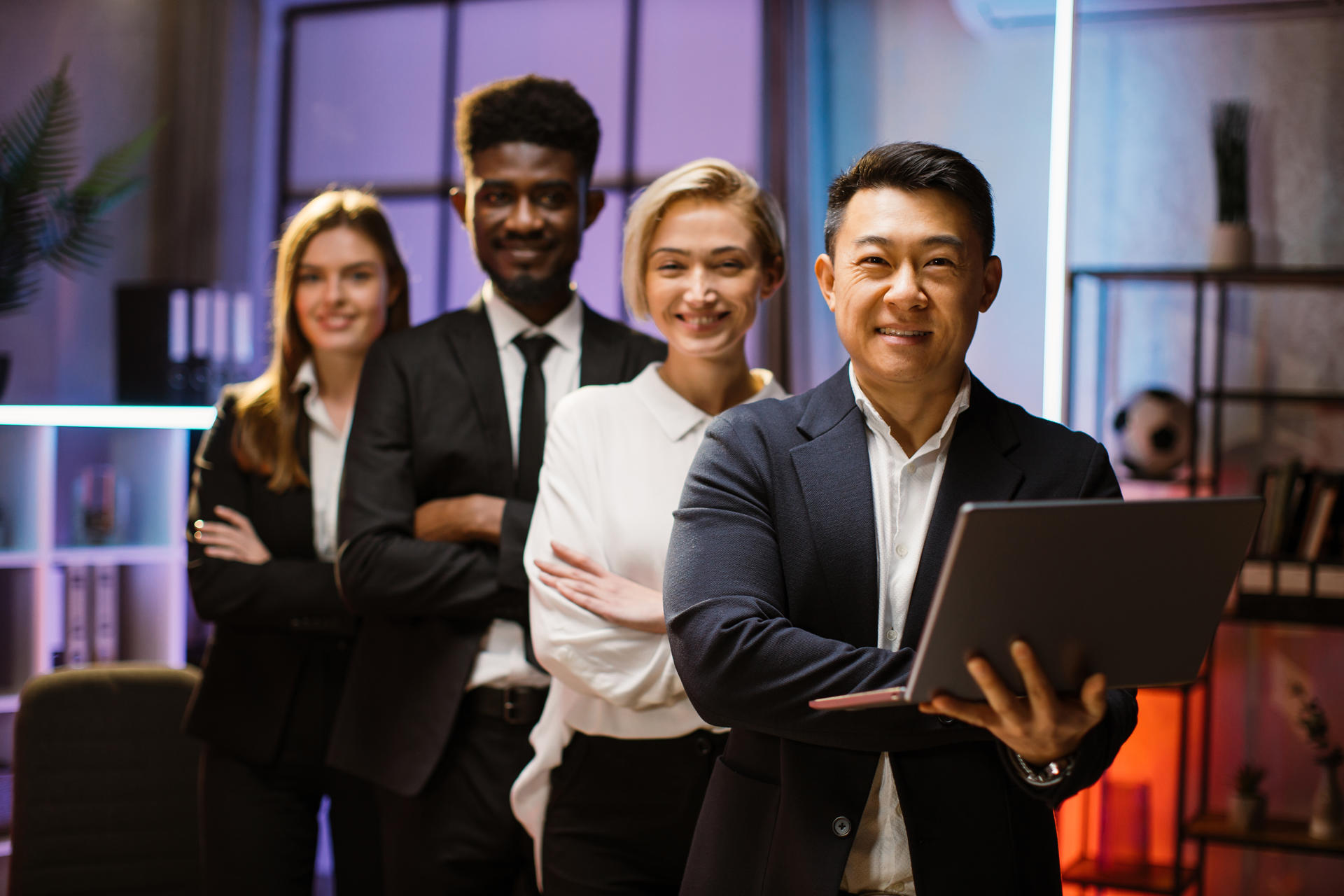 Happy multiethnic coworkers standing in a row in creative office, posing on camera with arms crossed. Confident male Asian ceo with laptop standing in front of his team