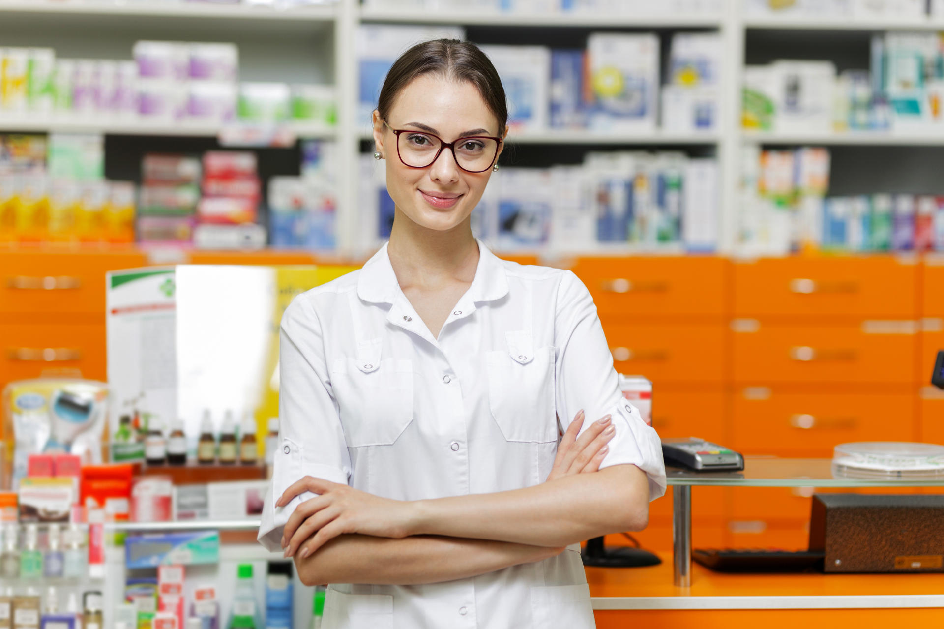 A pretty young girl with dark hair and glasses,wearing a medical overall,stands by the cash desk with her arms crossed  in a up-to-date pharmacy. .