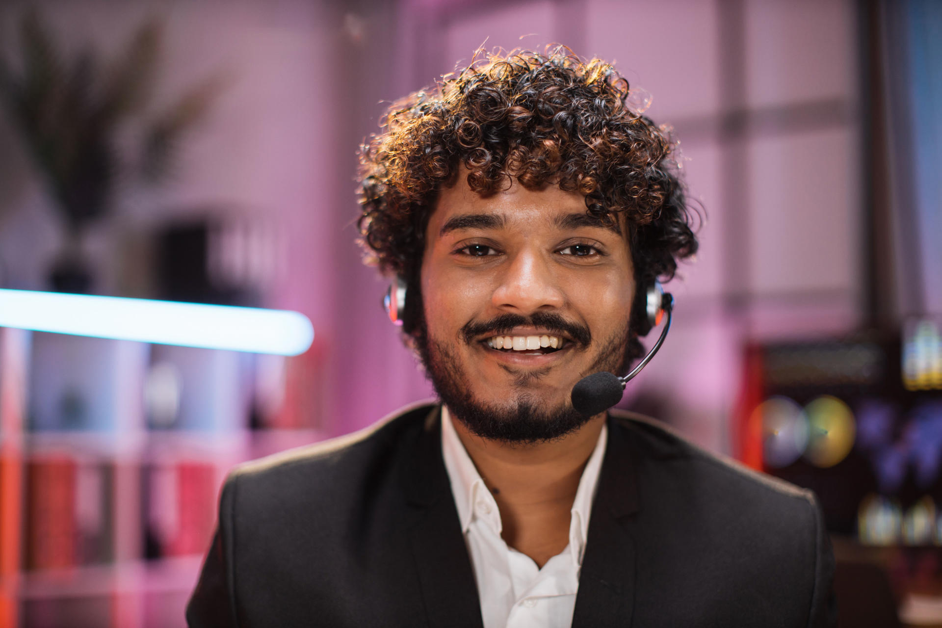 Smiling positive confident man in formal wear and headset sitting at office desk. Company worker with beard looking at camera. Concept of people and business.
