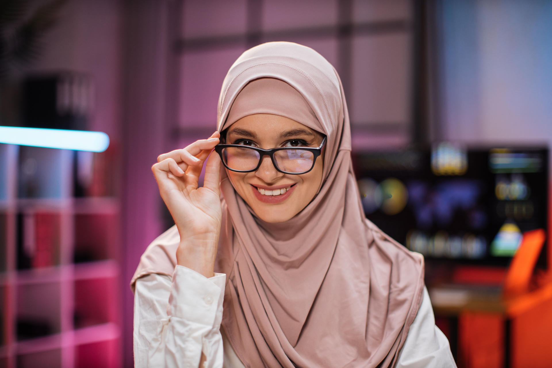 Smiling positive confident woman in formal wear and hijab and eyeglasses sitting at office desk. Company worker with headscarf looking at camera. Concept of people and business.