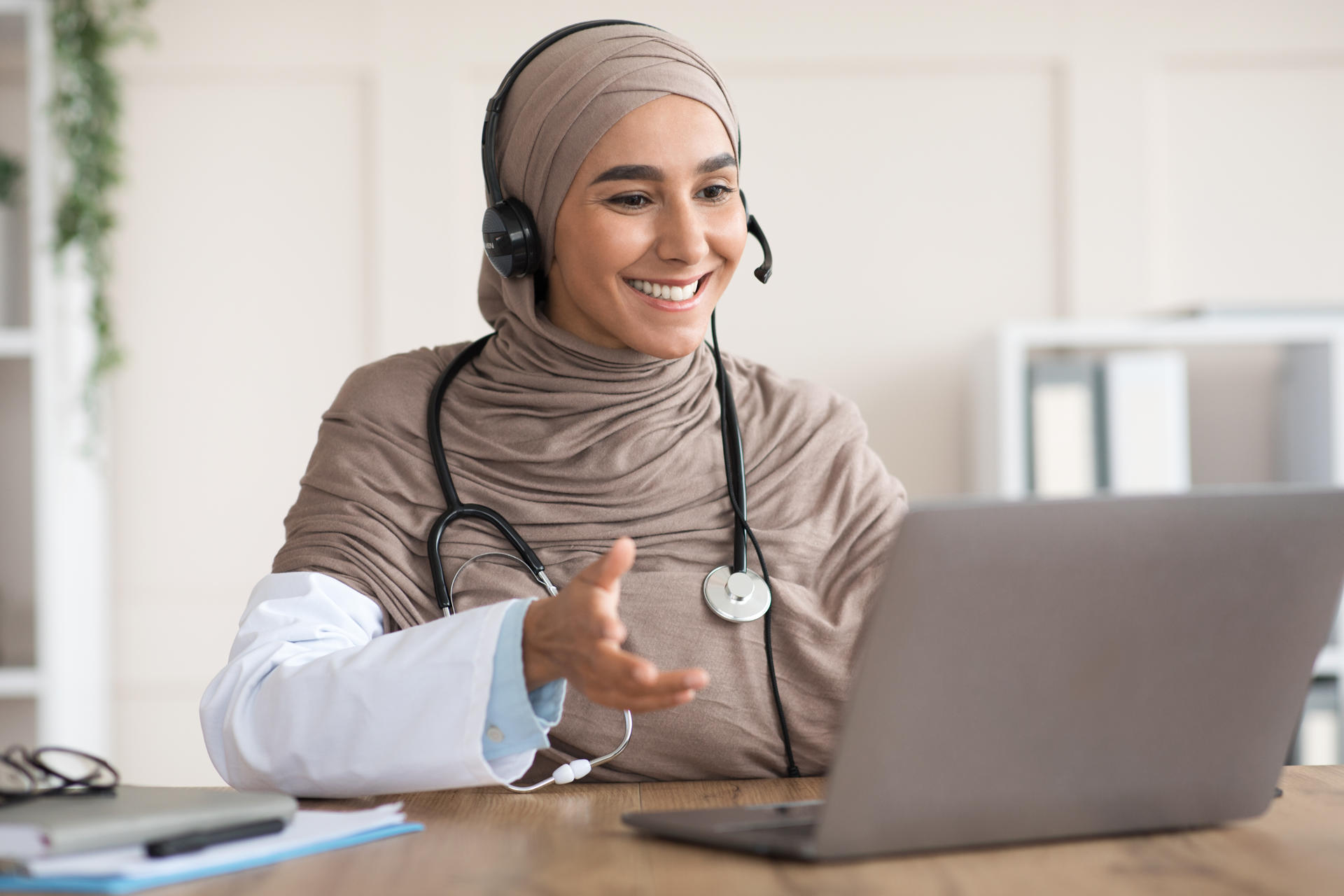 Cheerful arab female doctor working online in the clinic, using headset and laptop. Muslim woman therapist having video conference with her patient during isolation, hospital interior, copy space
