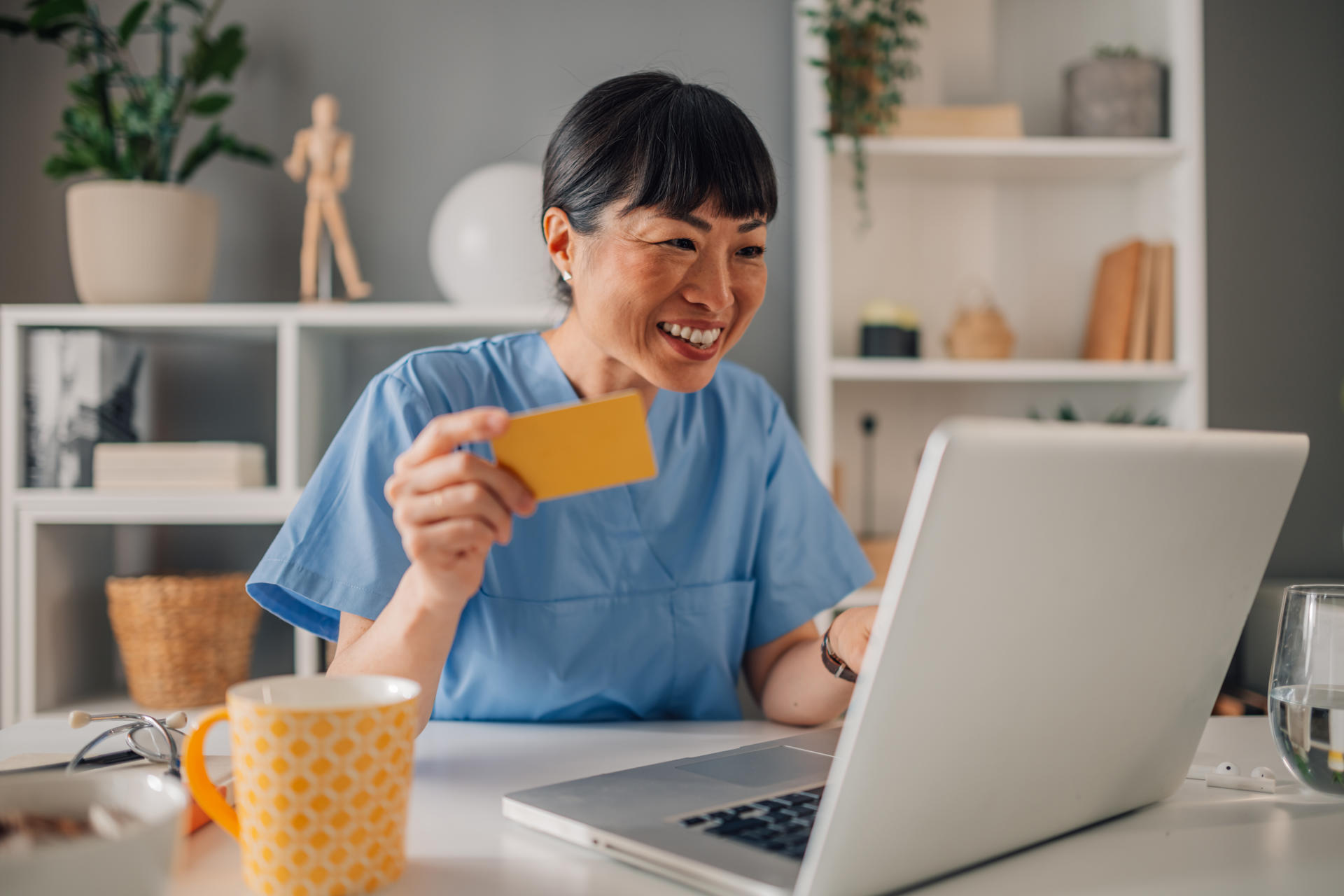 Asian female healthcare worker dressed in blue scrubs holds a yellow card while looking pensive at her laptop. She sits in a clean, organized, and thoughtfully decorated office space.