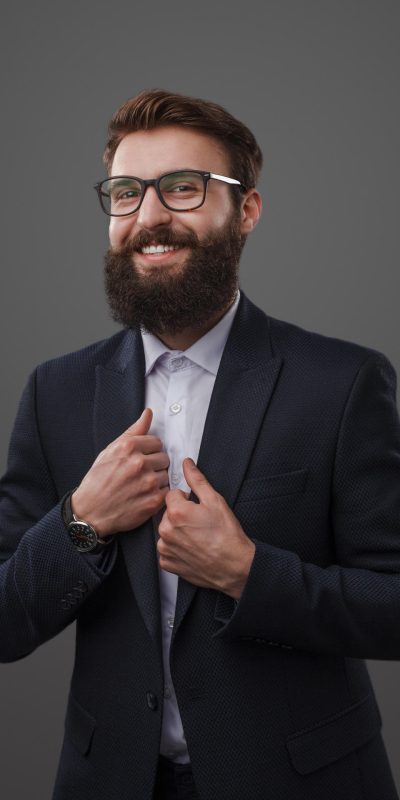 Handsome cheerful man wearing suit Classy male model with beard wearing elegant black suit with glasses and posing with smile at camera on gray.