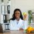 African American female dietitian holding apple and smiling at camera in modern office African American female dietitian holding apple and smiling at camera in modern office. Cheerful black nutritionist with laptop offering online consultations at weight loss clinic